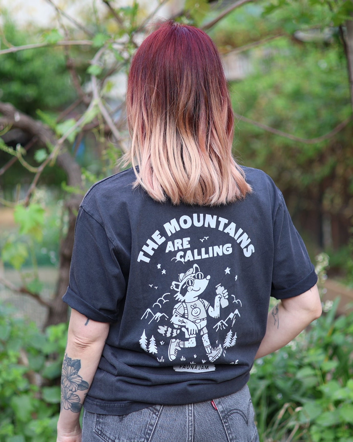 Person wearing a black t-shirt with 'The Mountains Are Calling' text and graphic, standing outdoors.