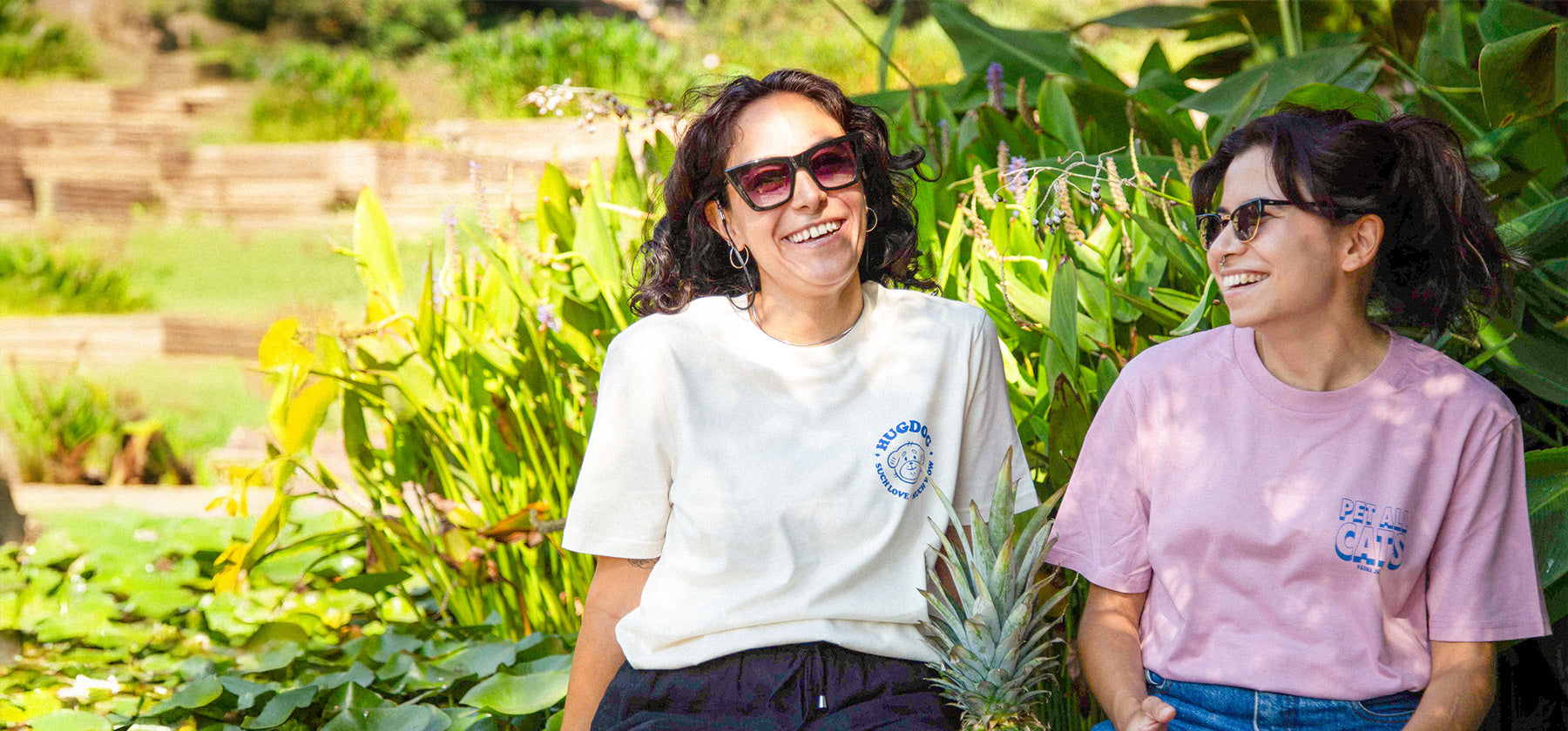 Two women sitting outdoors among greenery, wearing sunglasses.