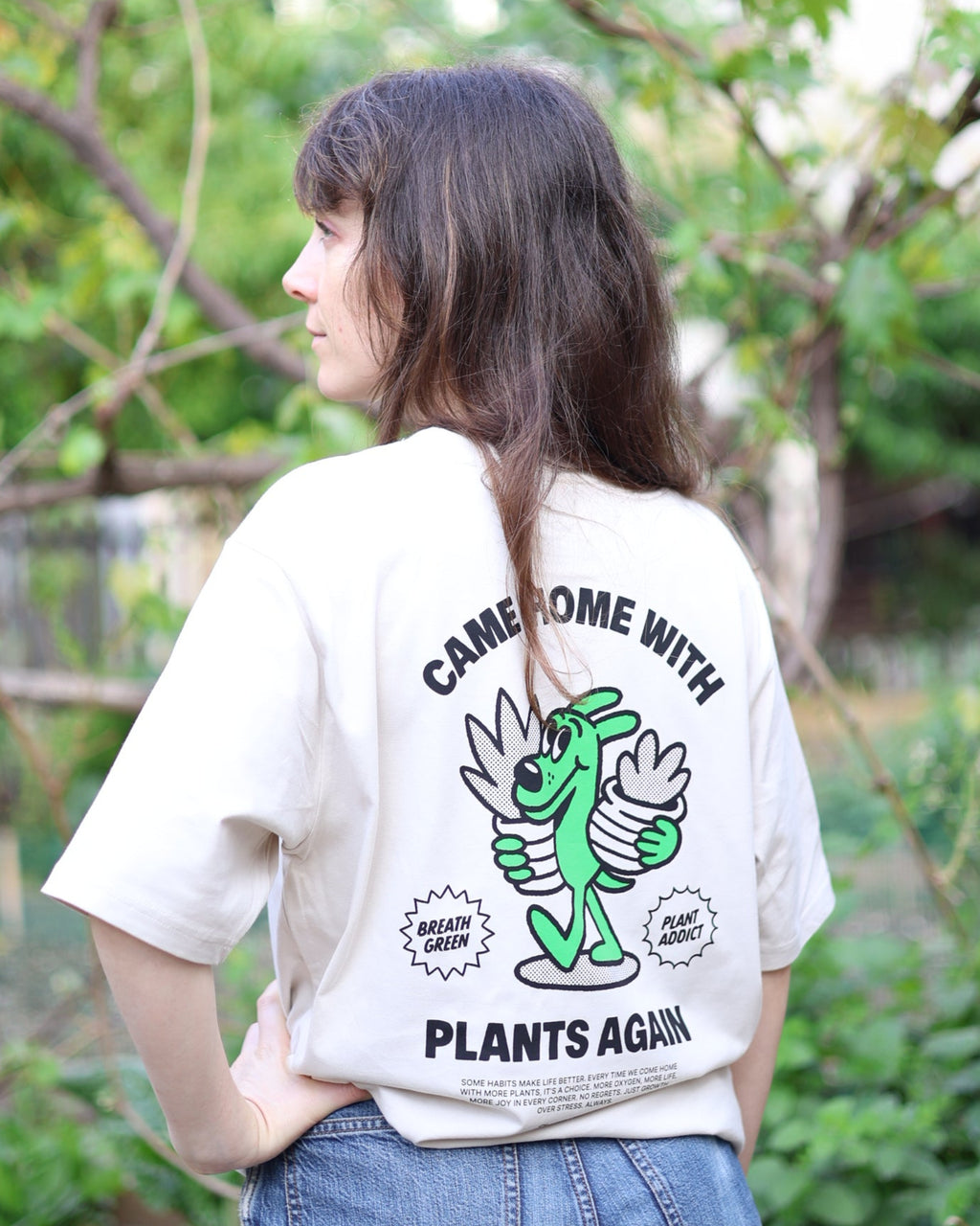 Person wearing a white t-shirt with green graphics and text, standing outdoors with greenery in the background