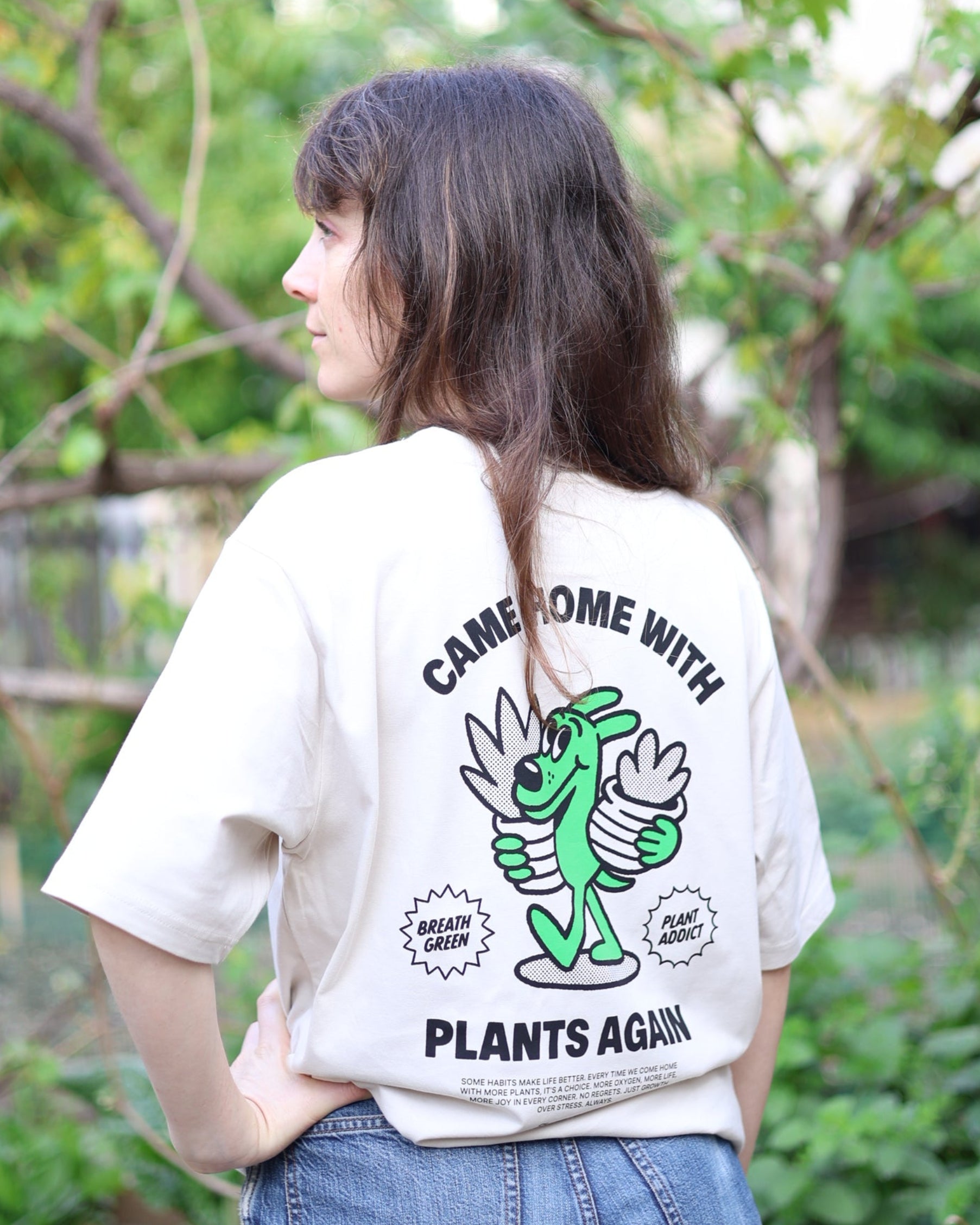 Person wearing a white t-shirt with green graphics and text, standing outdoors with greenery in the background
